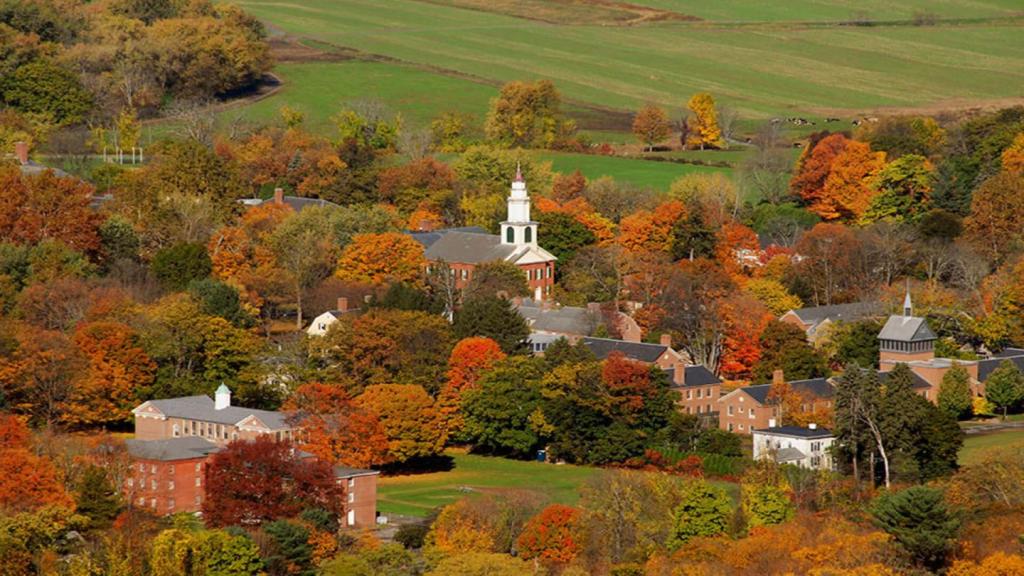 Aerial view of a picturesque town surrounded by vibrant autumn foliage, featuring historic buildings and a church steeple amidst a landscape of rolling hills.